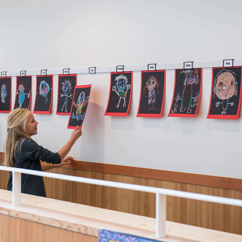 Leerling hangt een zelfgemaakte tekening op aan een Artiteq Info Rail in een basisschool, voor een kleurrijke en flexibele aankleding van het klaslokaal